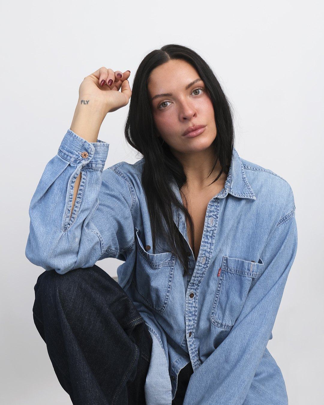 Rachel in an oversized denim shirt, long dark hair, hand resting at her temple